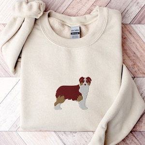 May include: Cream-colored sweatshirt with an embroidered image of a dog. The dog is depicted in shades of brown, white, and tan. The sweatshirt is folded, with the brand label visible.