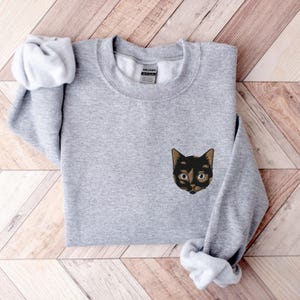 Op de afbeelding: Een grijs sweatshirt met een geborduurd zwart kattengezicht op de voorkant. De kat heeft bruin haar rond zijn ogen en een zwarte neus.