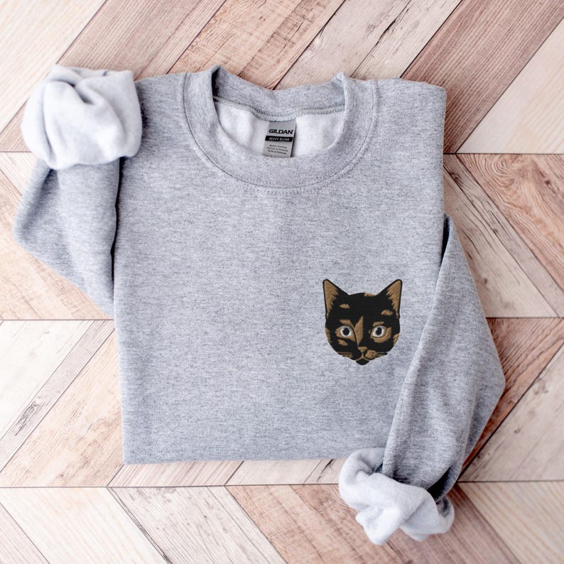 Op de afbeelding: Een grijs sweatshirt met een geborduurd zwart kattengezicht op de voorkant. De kat heeft bruin haar rond zijn ogen en een zwarte neus.