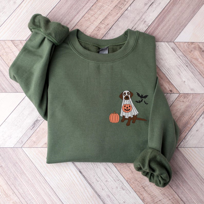 May include: A green sweatshirt featuring a brown dog dressed as a ghost, holding a pumpkin. The dog is standing next to a pumpkin and there are bats flying above it.