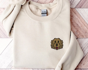 May include: A beige sweatshirt with a brown and black embroidered dog face on the left chest. The sweatshirt has a crew neck and long sleeves. The tag on the sweatshirt reads "Gildan Soft Style".
