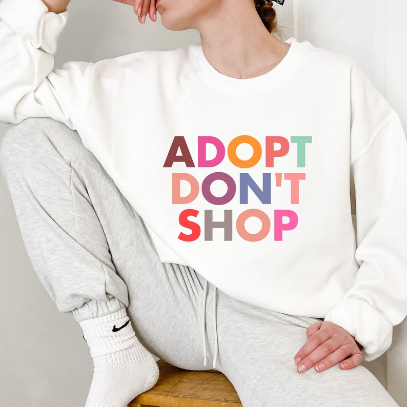 May include: White sweatshirt with the text "ADOPT DON'T SHOP" in colorful letters.
