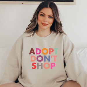May include: A beige sweatshirt with the text "ADOPT DON'T SHOP" in colorful letters. The letters are in different colors, including red, orange, yellow, green, blue, purple, pink, and brown.