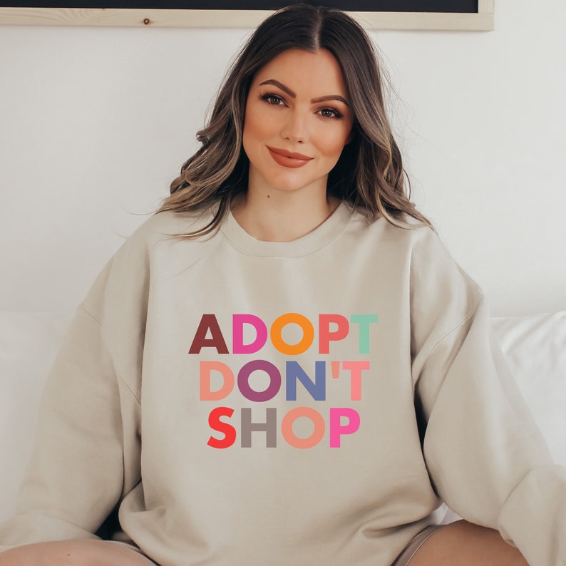 May include: A beige sweatshirt with the text "ADOPT DON'T SHOP" in colorful letters. The letters are in different colors, including red, orange, yellow, green, blue, purple, pink, and brown.