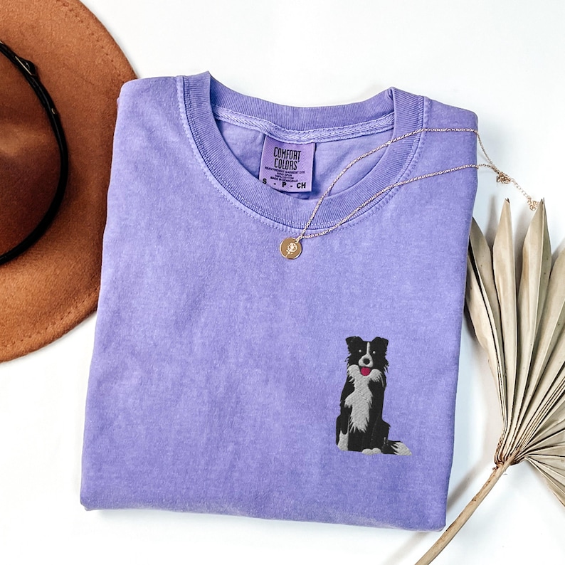 May include: A light purple Comfort Colors t-shirt with a small embroidered black and white Border Collie dog. A gold necklace with a pendant is draped across the shirt. The shirt has a crew neck and the Comfort Colors label is visible.