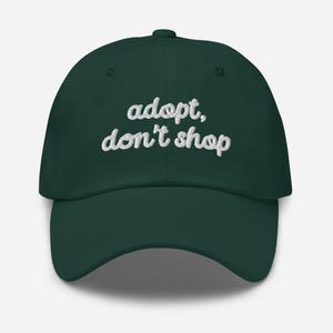 May include: A dark green baseball cap with the embroidered text "adopt, don't shop" in white cursive lettering. The cap has a curved brim and a button on top.