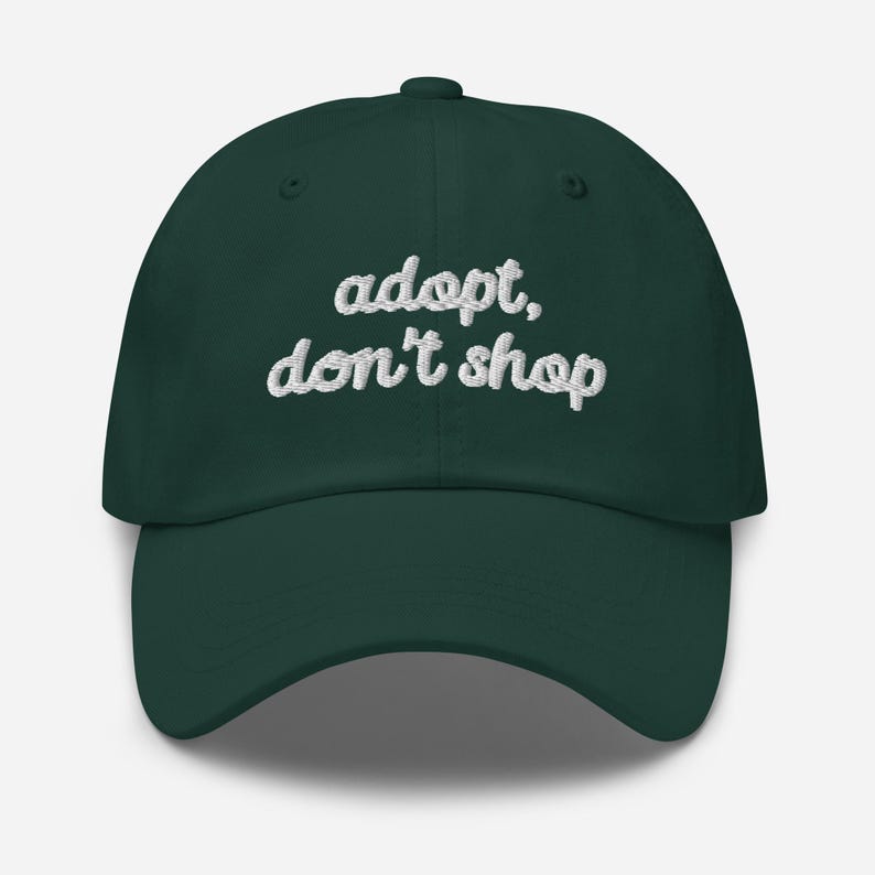 May include: A dark green baseball cap with the embroidered text "adopt, don't shop" in white cursive lettering. The cap has a curved brim and a button on top.