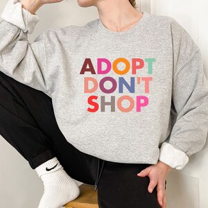 May include: A gray sweatshirt with the text "ADOPT DON'T SHOP" in colorful letters. The letters are in different colors, including red, orange, pink, purple, blue, and green.