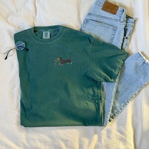 May include: A green t-shirt with a dachshund graphic holding flowers, paired with light blue jeans and round eyeglasses. The shirt is a solid color, and the jeans have a leather patch. The items are arranged on a white surface.