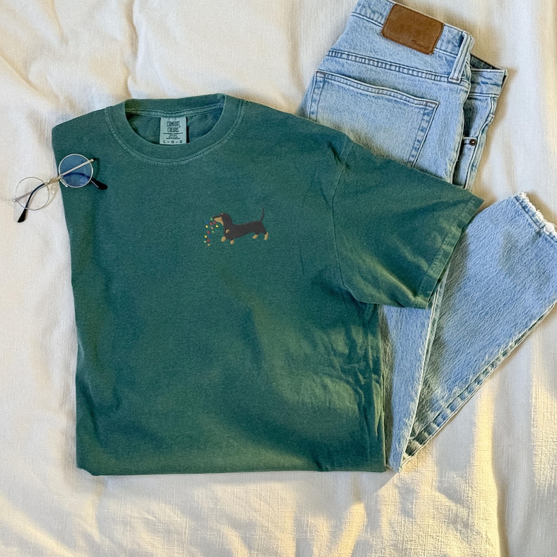 May include: A green t-shirt with a dachshund graphic holding flowers, paired with light blue jeans and round eyeglasses. The shirt is a solid color, and the jeans have a leather patch. The items are arranged on a white surface.