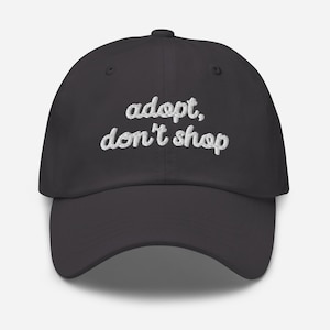 May include: A dark gray baseball cap with the embroidered text "adopt, don't shop" in white cursive lettering. The cap has a curved brim and a button on top. This hat is a stylish accessory for animal lovers.