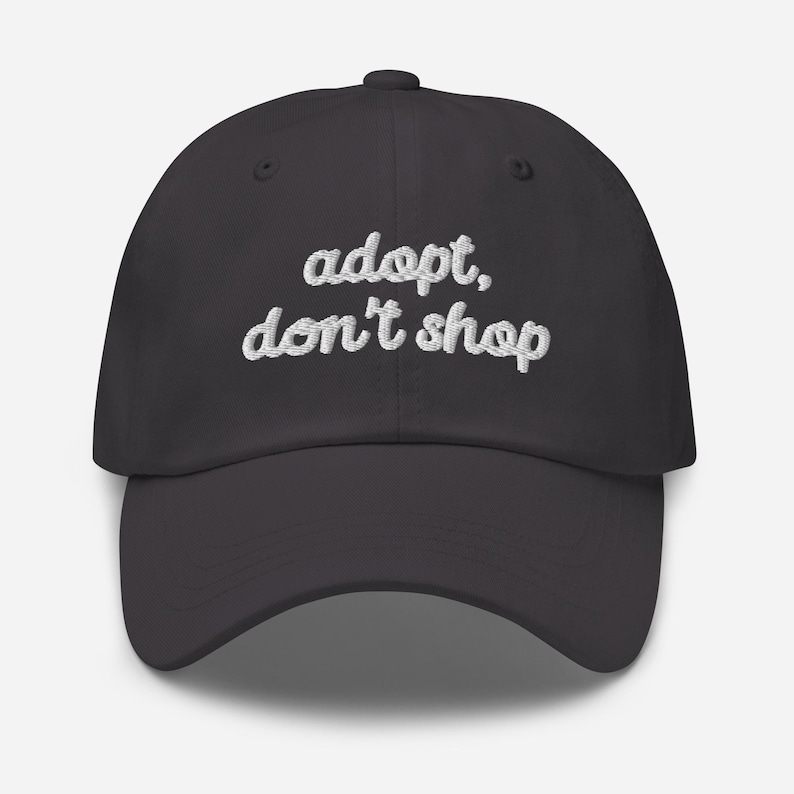 May include: A dark gray baseball cap with the embroidered text "adopt, don't shop" in white cursive lettering. The cap has a curved brim and a button on top. This hat is a stylish accessory for animal lovers.
