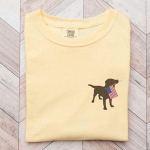 Puede incluir: Una camiseta amarilla con una silueta marr&oacute;n de un perro que sostiene una bandera estadounidense en su boca. La camiseta tiene una peque&ntilde;a etiqueta que dice "Comfort Colors".