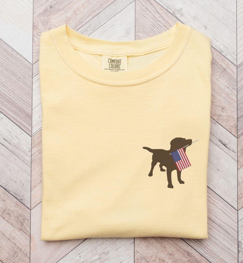 Puede incluir: Una camiseta amarilla con una silueta marr&oacute;n de un perro que sostiene una bandera estadounidense en su boca. La camiseta tiene una peque&ntilde;a etiqueta que dice "Comfort Colors".