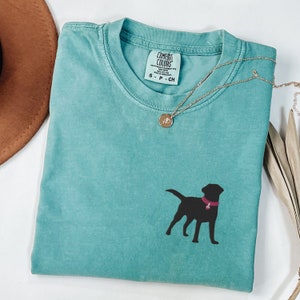 May include: A teal blue t-shirt with a black silhouette of a labrador retriever wearing a pink collar. The shirt has a tag that reads "Comfort Colors" and "S - P - CH".