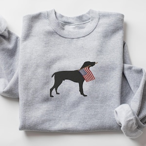 May include: A gray sweatshirt with a black silhouette of a dog carrying an American flag in its mouth.