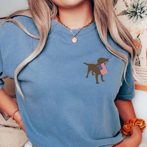Puede incluir: Una camiseta azul con una silueta marr&oacute;n de un perro sosteniendo una bandera estadounidense.