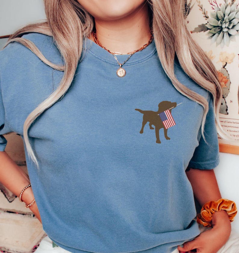 Puede incluir: Una camiseta azul con una silueta marr&oacute;n de un perro sosteniendo una bandera estadounidense.