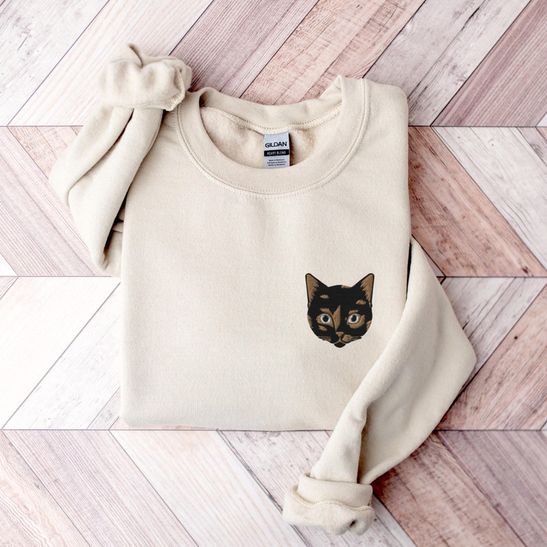 Embroidered Tortoiseshell Cat on Unisex Gildan 18000 Sweatshirt in Sand Color which is Beige in tone, folded