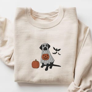 May include: A cream-colored sweatshirt with a black dog wearing a ghost costume and holding a jack-o'-lantern. Two bats are flying above the dog. The dog is holding a pumpkin.