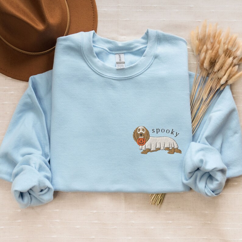 May include: Light blue sweatshirt with a cartoon dachshund wearing a ghost costume and holding a pumpkin. The text "spooky" is above the dog.