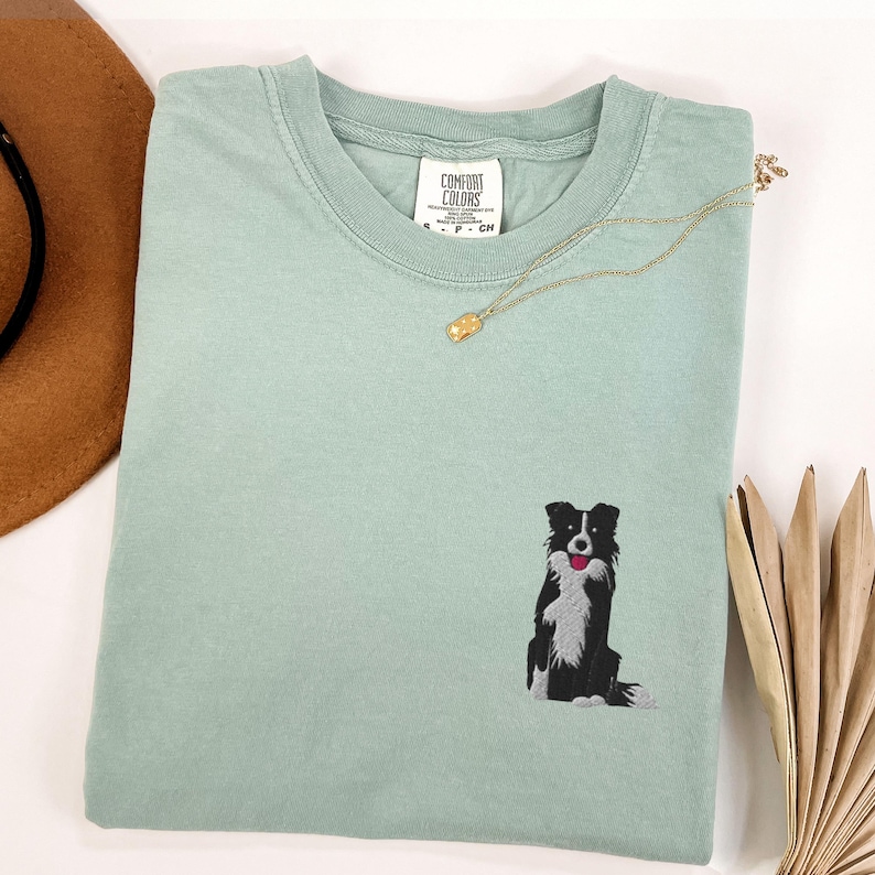 May include: A light green t-shirt with a small embroidered black and white dog design. The shirt has a crew neck and a tag that reads "Comfort Colors". A gold necklace with a small pendant is draped across the shirt.