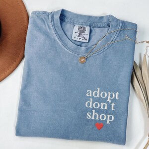 May include: A light blue t-shirt with the text "adopt don't shop" in white embroidery and a small red heart below the text. The shirt is made of a soft, comfortable material.