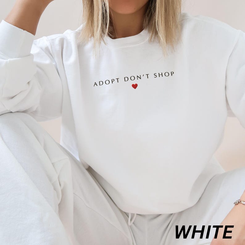 May include: White sweatshirt featuring the text "ADOPT DON'T SHOP" in black, with a small red heart. The word "WHITE" is at the bottom of the image. The sweatshirt is part of a matching set.