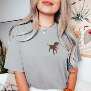 Puede incluir: Una camiseta gris con una silueta marr&oacute;n de un perro sosteniendo una peque&ntilde;a bandera estadounidense.