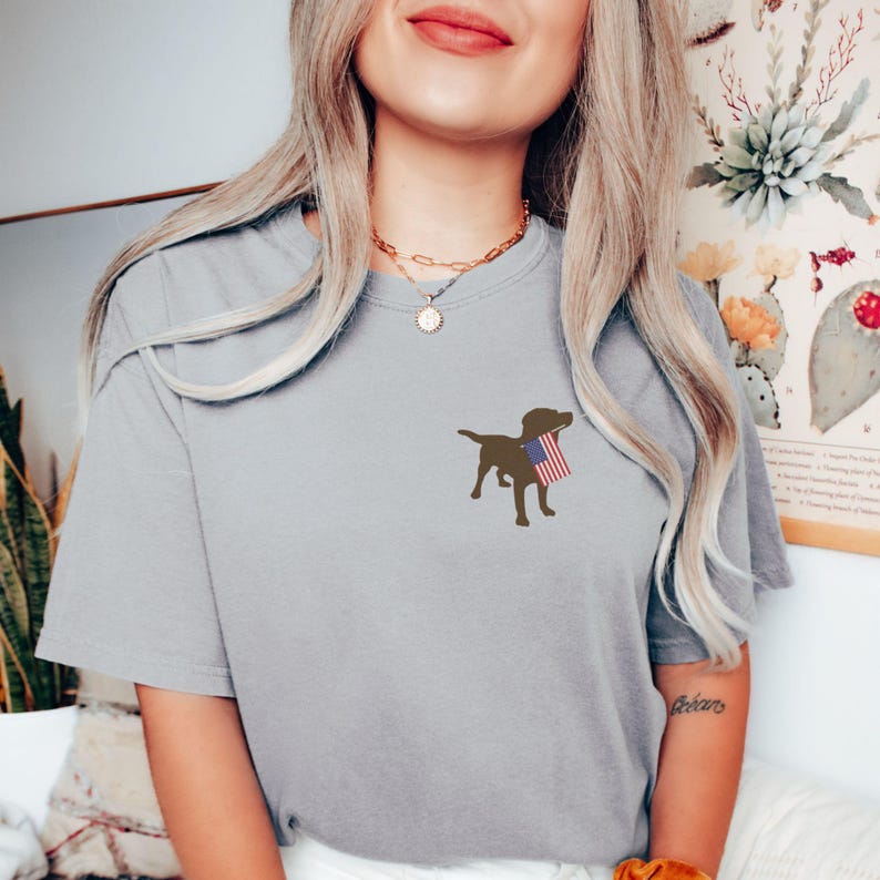 Puede incluir: Una camiseta gris con una silueta marr&oacute;n de un perro sosteniendo una peque&ntilde;a bandera estadounidense.
