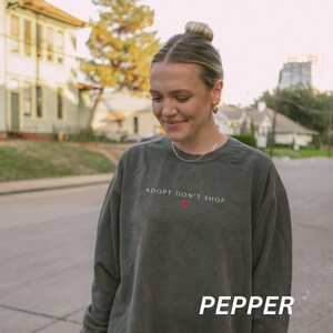 May include: A dark green sweatshirt with the text "ADOPT DON'T SHOP" in white and a red heart. The word "PEPPER" is printed in white at the bottom. The sweatshirt is worn by a person.