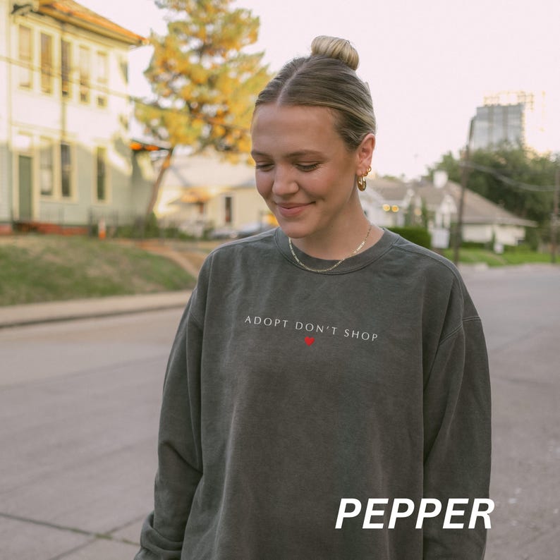 May include: A dark green sweatshirt with the text "ADOPT DON'T SHOP" in white and a red heart. The word "PEPPER" is printed in white at the bottom. The sweatshirt is worn by a person.