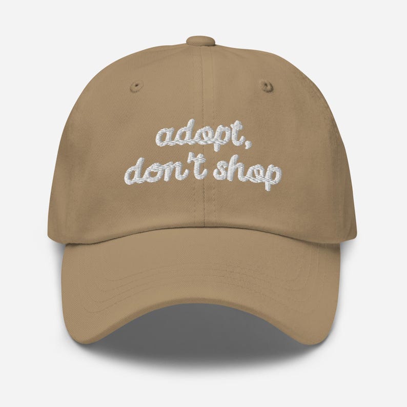 May include: A tan baseball cap with the embroidered text "adopt, don't shop" in white. The cap has a curved brim and a button on top. This hat is a stylish accessory for animal lovers.