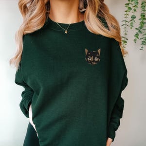 Op de afbeelding: Een donkergroen sweatshirt met een geborduurd zwart kattengezicht op de linkerborst. De kat heeft bruine ogen en een witte neus.