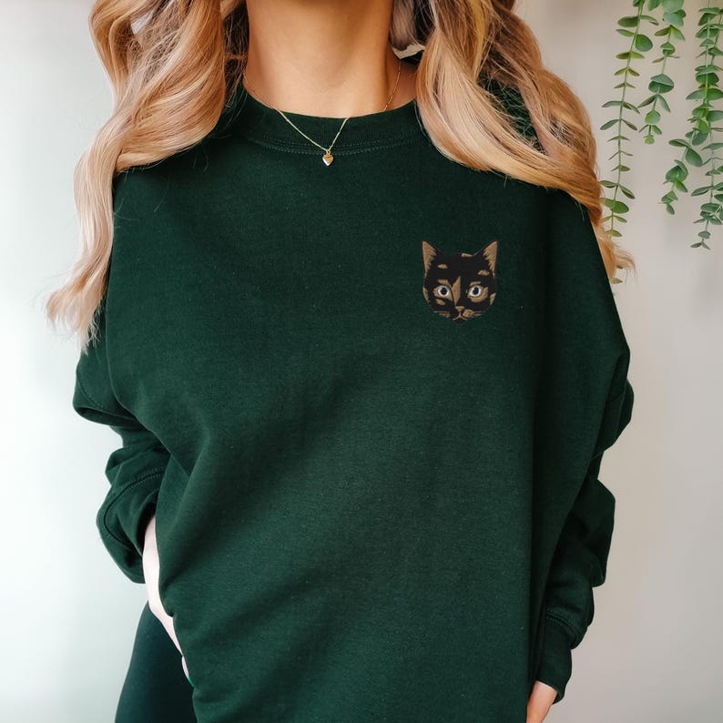 Op de afbeelding: Een donkergroen sweatshirt met een geborduurd zwart kattengezicht op de linkerborst. De kat heeft bruine ogen en een witte neus.