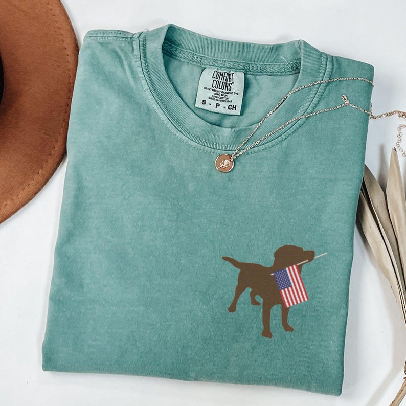 Puede incluir: Una camiseta azul turquesa con una silueta marr&oacute;n de un perro sosteniendo una peque&ntilde;a bandera estadounidense. La camiseta tiene una etiqueta que dice "Comfort Colors" y "S - P - CH".