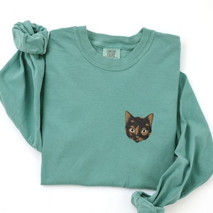 May include: A teal long-sleeved t-shirt with a black cat face embroidered on the front. The shirt has a crew neck and the text "Comfort Colors" on a tag.