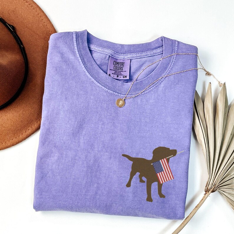 Puede incluir: Una camiseta morada con una silueta marr&oacute;n de un perro sosteniendo una bandera estadounidense. La camiseta est&aacute; hecha por Comfort Colors.