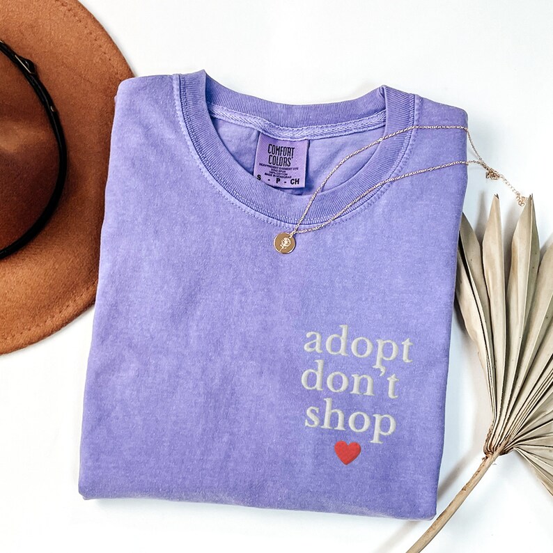 May include: Purple t-shirt with the text "adopt don't shop" and a red heart. The shirt is made by Comfort Colors.