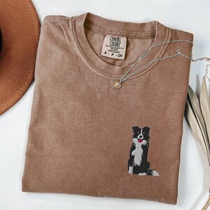 May include: A brown Comfort Colors t-shirt with a small embroidered black and white dog graphic. A silver necklace with a pendant rests on the shirt. The shirt has a crew neck and short sleeves.