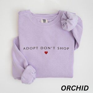 May include: A lilac sweatshirt featuring the slogan "ADOPT DON'T SHOP" with a red heart. The word "ORCHID" is printed in a bold font at the bottom. The sweatshirt has long sleeves and is made of a soft material.
