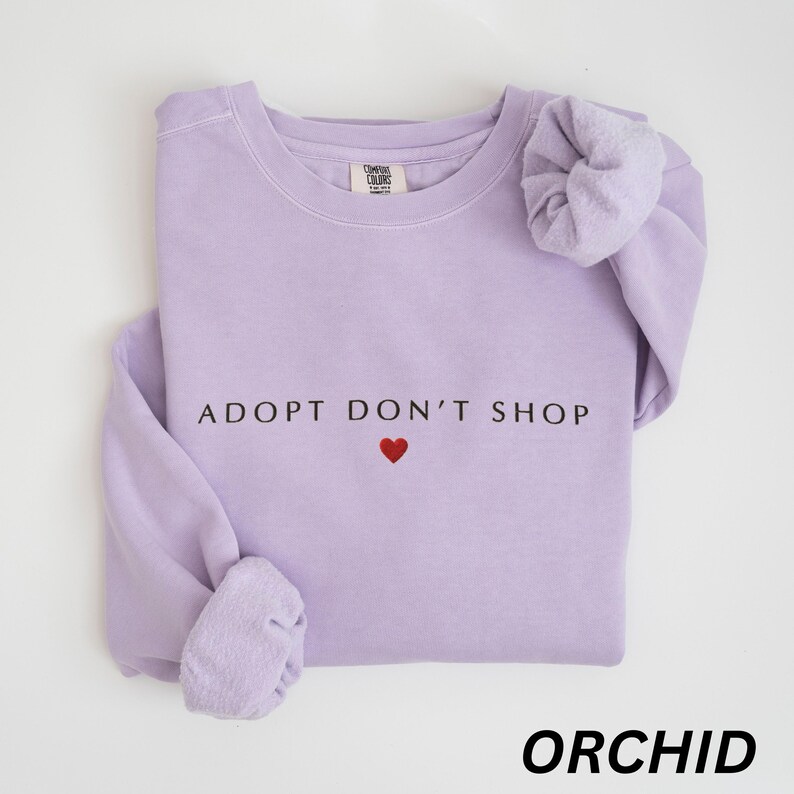May include: A lilac sweatshirt featuring the slogan "ADOPT DON'T SHOP" with a red heart. The word "ORCHID" is printed in a bold font at the bottom. The sweatshirt has long sleeves and is made of a soft material.