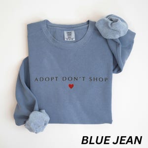 May include: A blue jean-coloured sweatshirt featuring the text "ADOPT DON'T SHOP" and a red heart. The sweatshirt has a crew neck and long sleeves. The words "BLUE JEAN" are printed at the bottom.