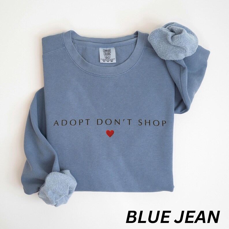 May include: A blue jean-coloured sweatshirt featuring the text "ADOPT DON'T SHOP" and a red heart. The sweatshirt has a crew neck and long sleeves. The words "BLUE JEAN" are printed at the bottom.
