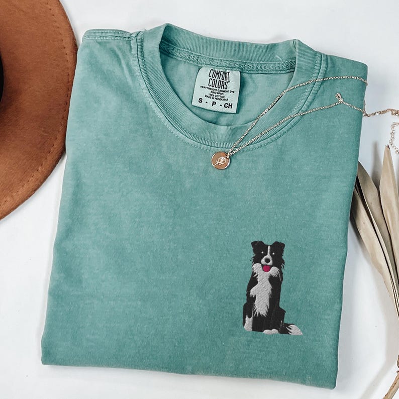 May include: A seafoam green Comfort Colors t-shirt featuring an embroidered black and white Border Collie dog. A delicate silver necklace with a pendant rests on the shirt. The shirt is folded on a white surface.