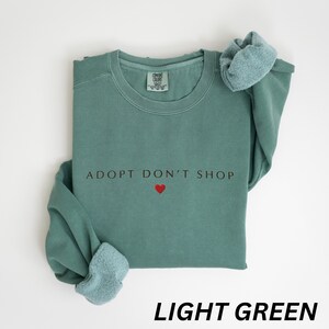 May include: A light green sweatshirt featuring the words "ADOPT DON'T SHOP" and a red heart. The garment has a crew neck and long sleeves. The words "LIGHT GREEN" are at the bottom of the image.