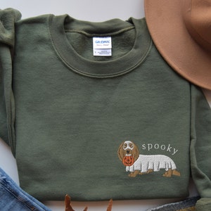 May include: A green sweatshirt with an embroidered image of a dachshund wearing a ghost costume and holding a jack-o-lantern. The text "spooky" is embroidered below the dog.