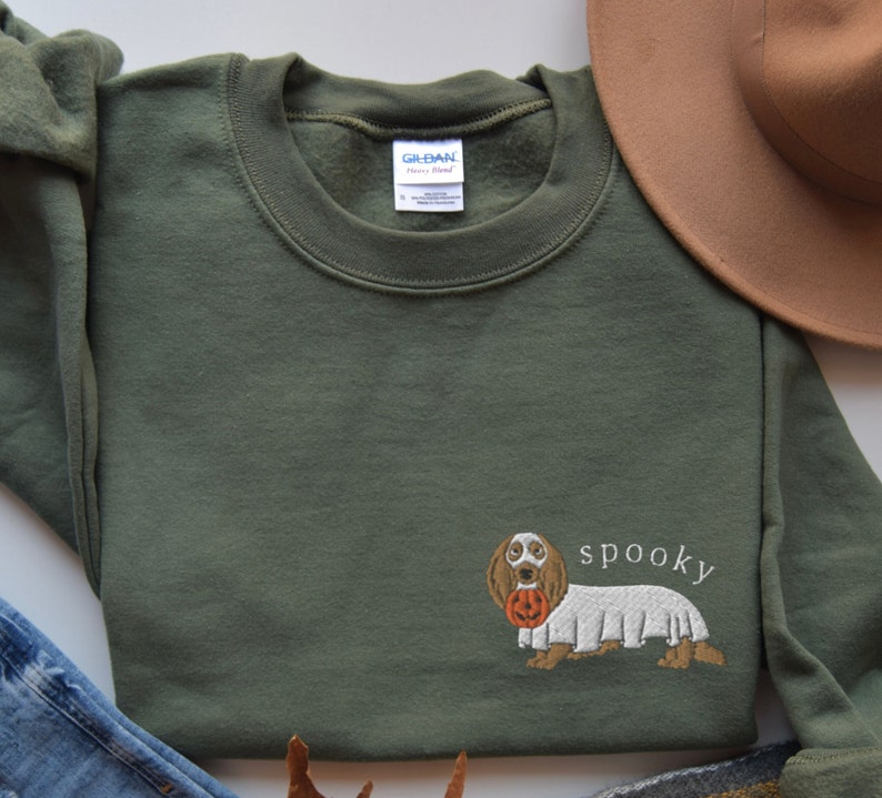 May include: A green sweatshirt with an embroidered image of a dachshund wearing a ghost costume and holding a jack-o-lantern. The text "spooky" is embroidered below the dog.