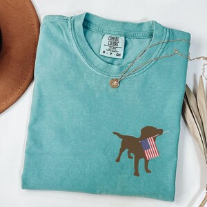 Puede incluir: Una camiseta azul turquesa con una silueta marr&oacute;n de un perro sosteniendo una bandera estadounidense. La camiseta tiene una etiqueta que dice "Comfort Colors" y "S - P - CH".