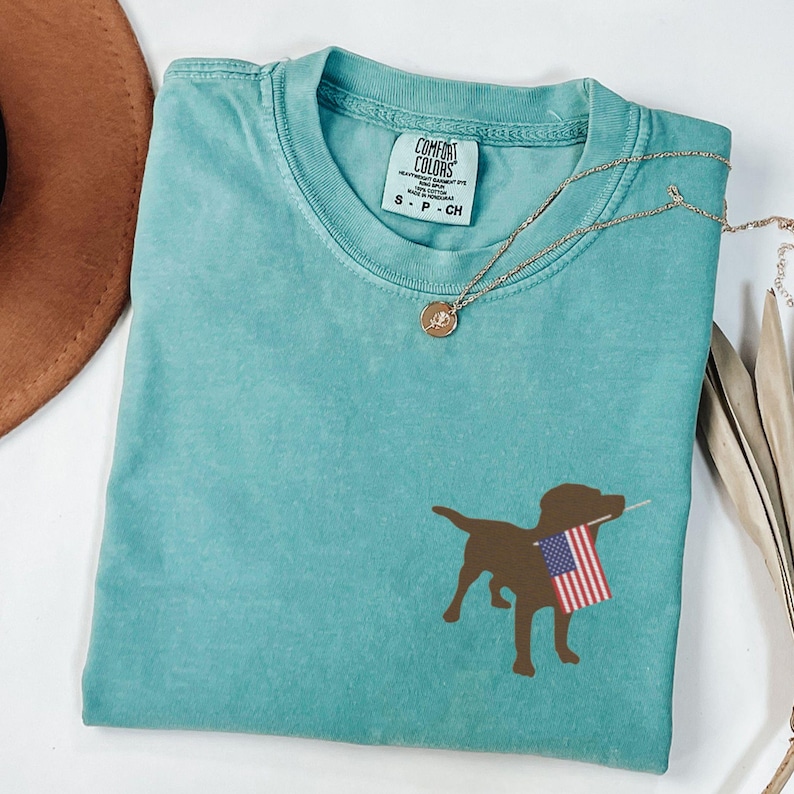 Puede incluir: Una camiseta azul turquesa con una silueta marr&oacute;n de un perro sosteniendo una bandera estadounidense. La camiseta tiene una etiqueta que dice "Comfort Colors" y "S - P - CH".